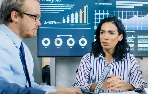 Man and woman conversating in front of a monitor displaying graphs and charts